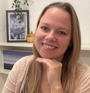 dreißig jährige Frau lächelt in die Kamera während sie an einem Schreibtisch vor ihrem Laptop sitzt, das Kinn auf einer Hand aufgestützt