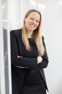 junge Frau mit blonden Haaren sieht lächelnd in die Kamera, hat ein schwarzes Kleid und einen schwarzen Blazer an